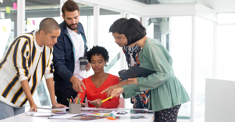 A group of individuals gathered around a table, with a man gesturing towards digital projects displayed on the surface.