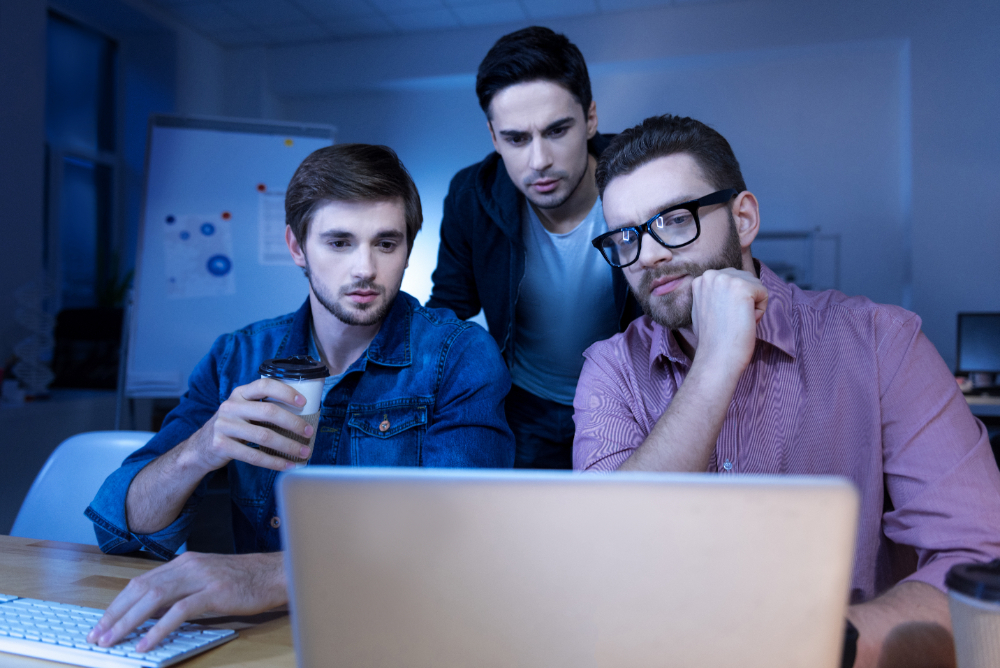 Three men collaborating on digital projects while examining a laptop computer together.