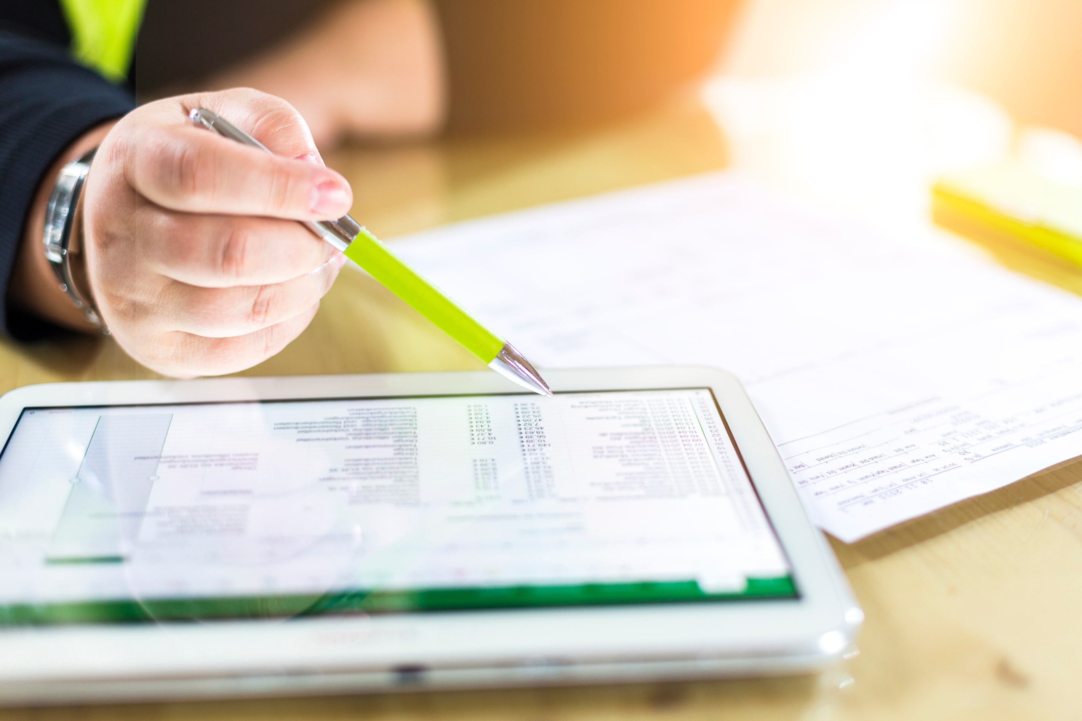 A person's hand grips a pen above a tablet, with paper in view, symbolizing custom CRM solutions.